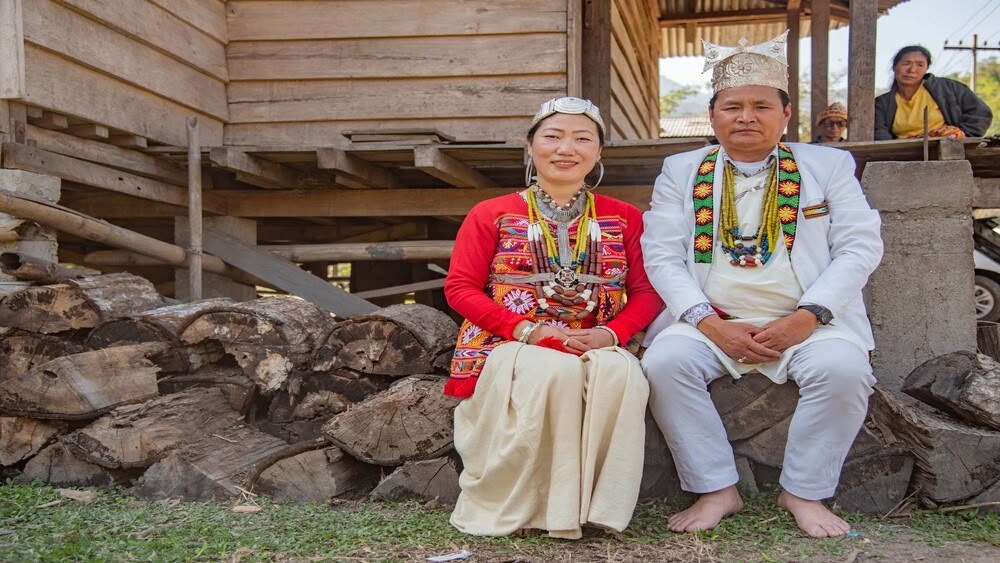 An Aka Hrusso couple in traditional red and white attire seated on a stone wall outside a wooden house