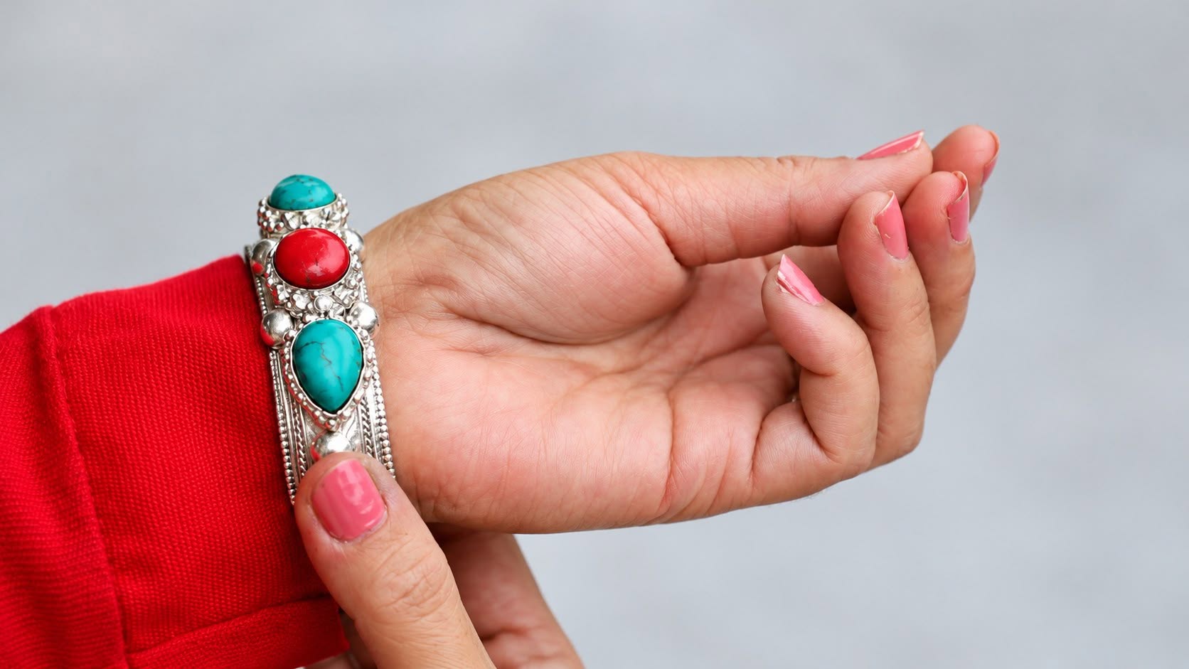 A hand wearing a silver ring set with turquoise and red stones, against a red sleeve
