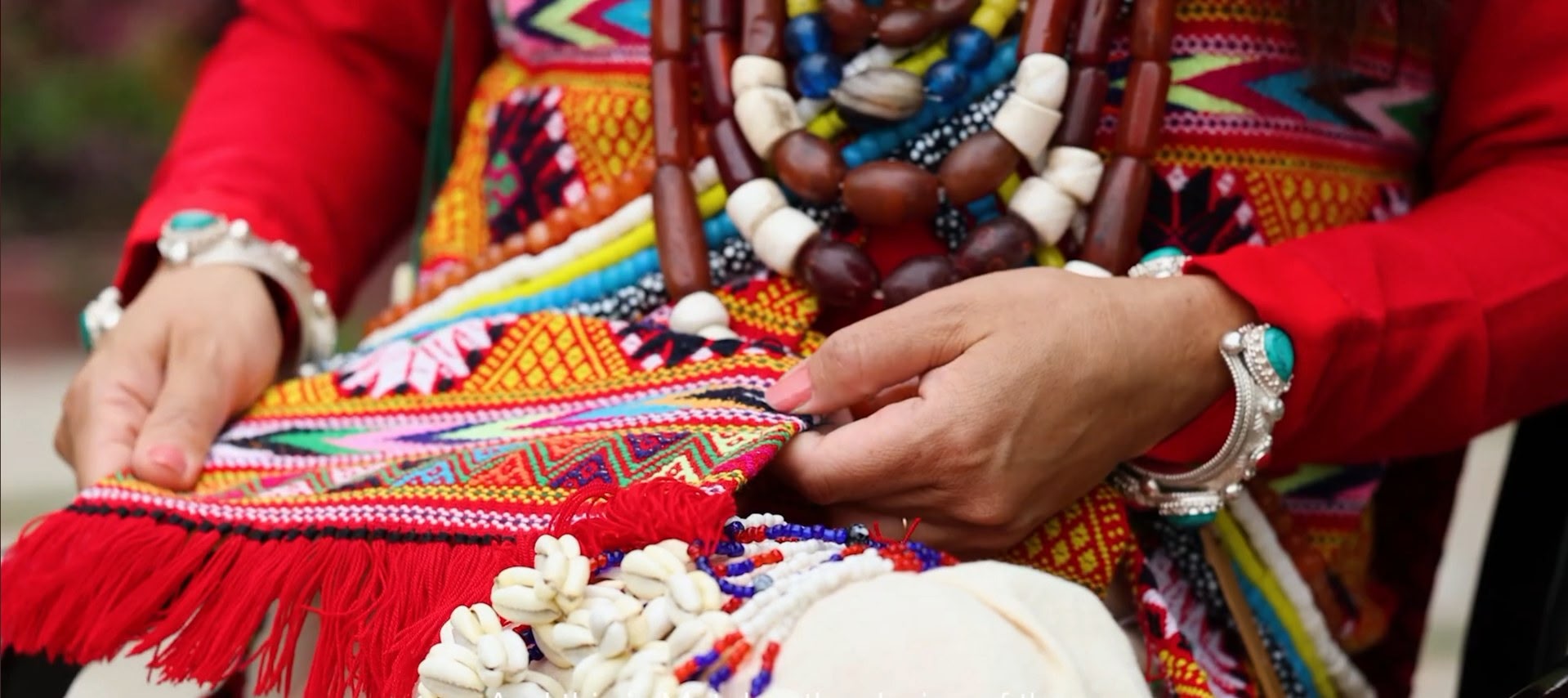 Close-up of hands holding a stone and bead necklace over a red woven Aka Hrusso garment