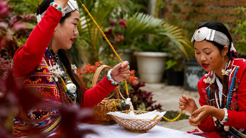 Two Aka Hrusso women in red garments and silver headbands stringing beads in a garden