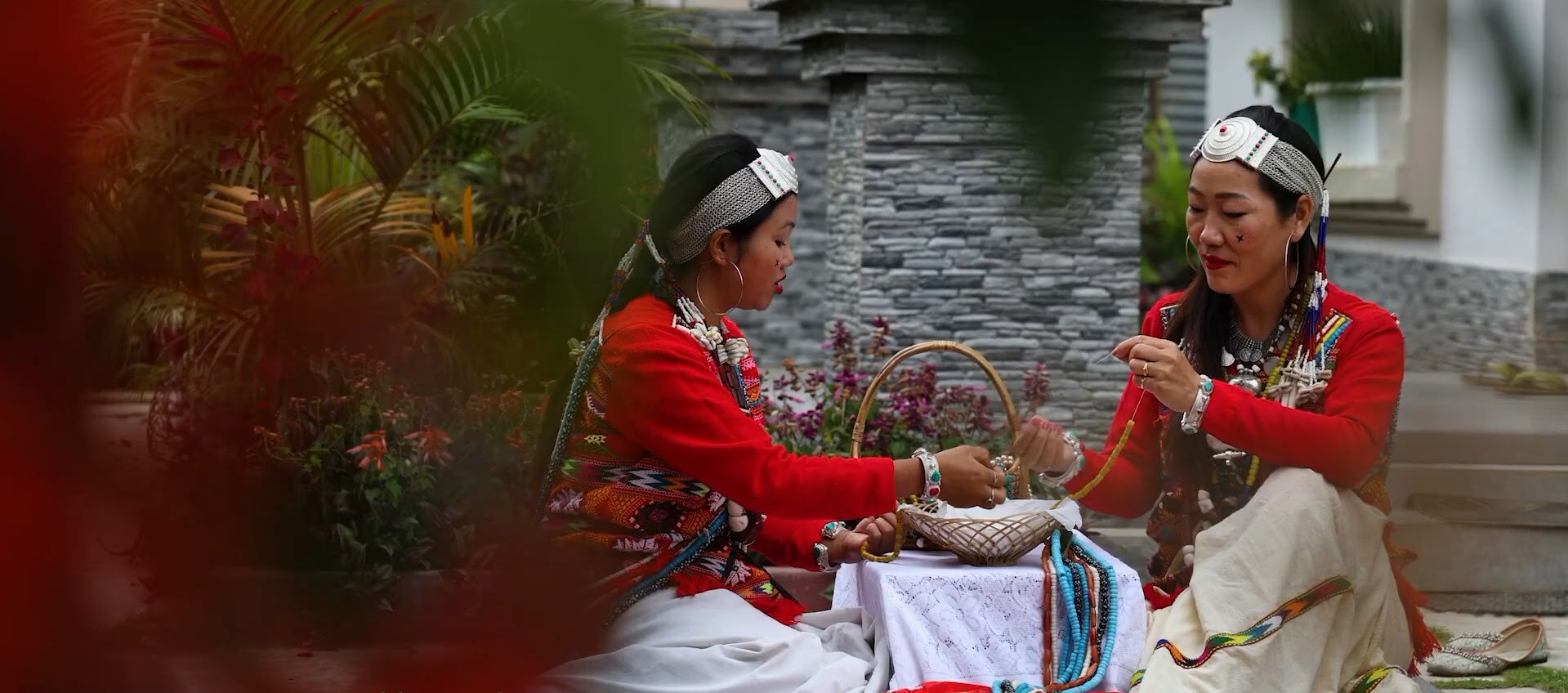 Two Aka Hrusso women seated at a table stringing beads in a courtyard