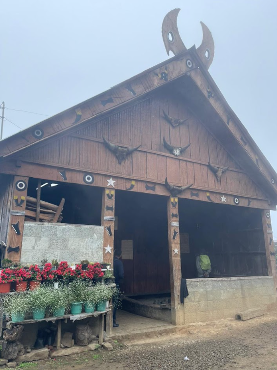 Wooden morung with carved horn roof and a row of flowering plants