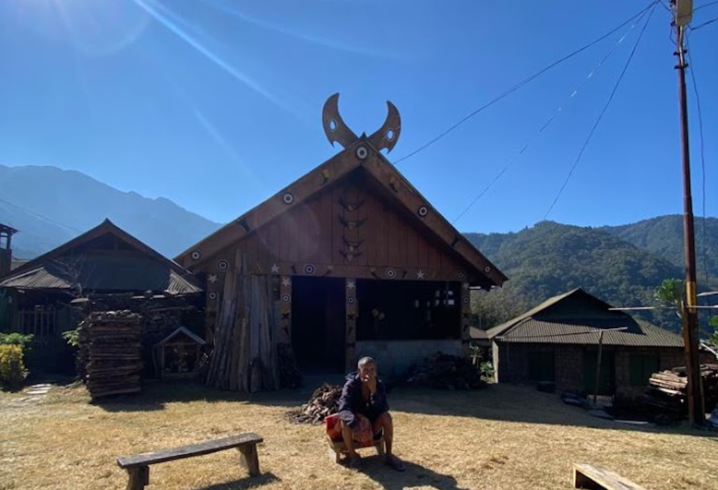 Angami wooden morung with horn-shaped roof and a man seated on a bench in front