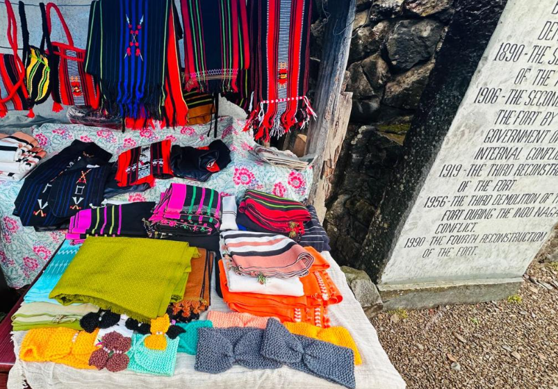Open-air honesty shop with colourful Naga textiles and bags displayed beside a stone memorial
