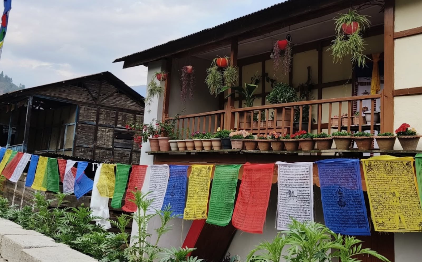 A line of colourful prayer flags strung in front of a wooden Himalayan house with flower pots on the balcony
