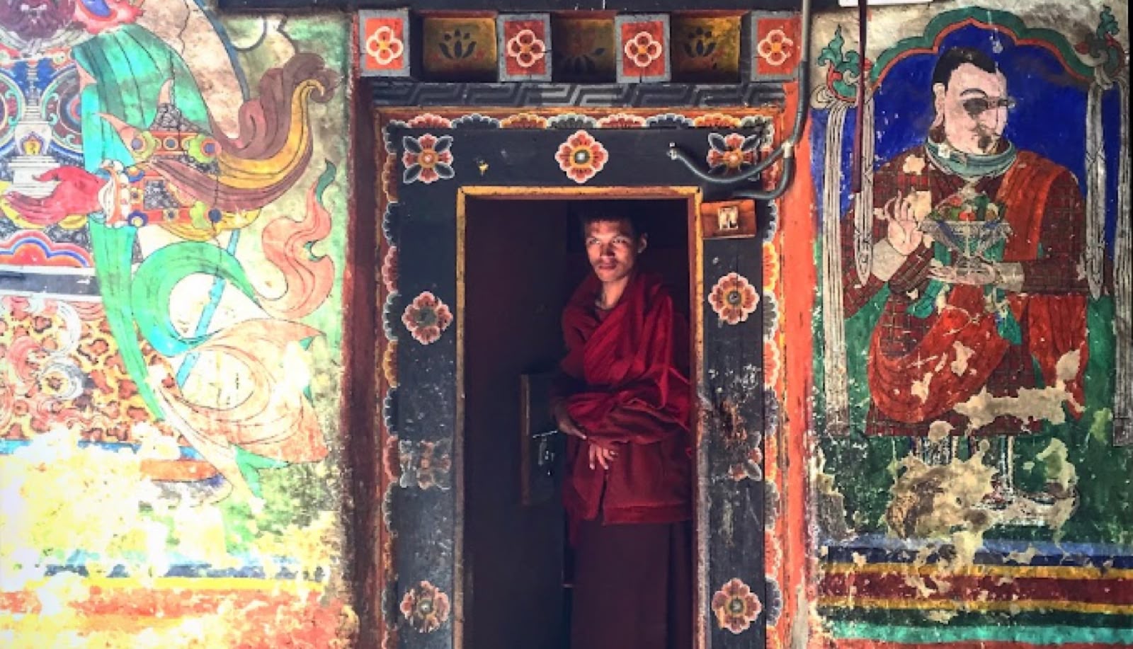 A young monk in maroon robes standing in a doorway framed by painted Buddhist murals