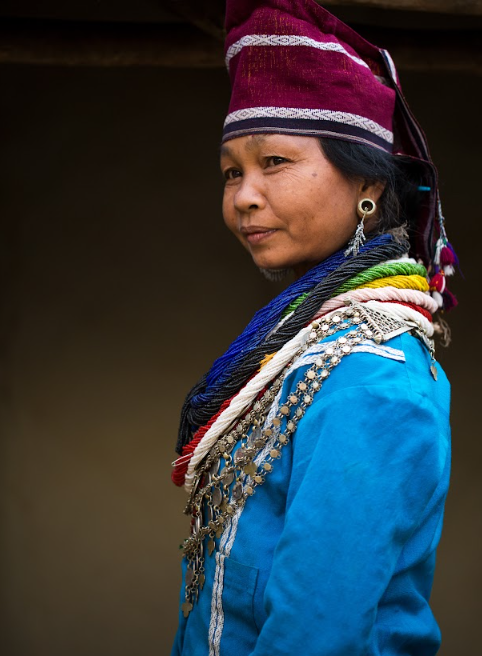 Side portrait of a Reang woman in a maroon cap and a blue top, wearing layered bead necklaces and a silver chain necklace