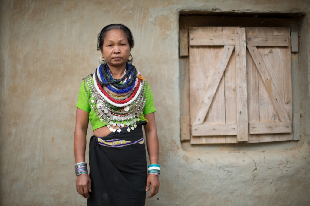 Portrait of a Reang woman standing against a clay wall beside a wooden shuttered window, wearing a green top, black wraparound skirt, layered bead necklaces with silver coins and silver bangles