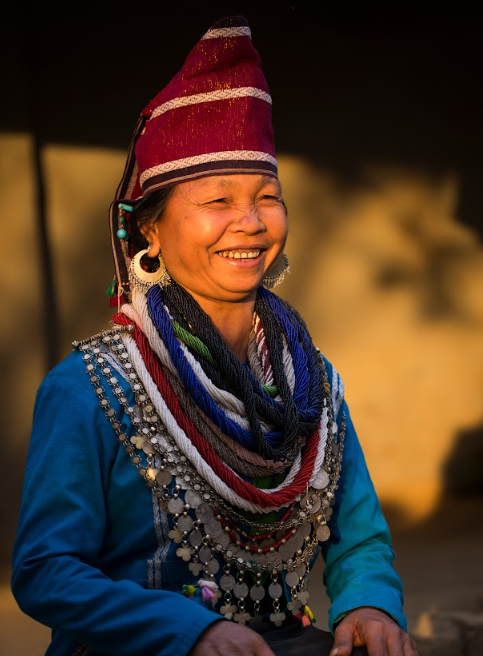 Smiling Reang woman in a red cap and blue top wearing hoop earrings, bead strands and a silver coin necklace in warm evening light