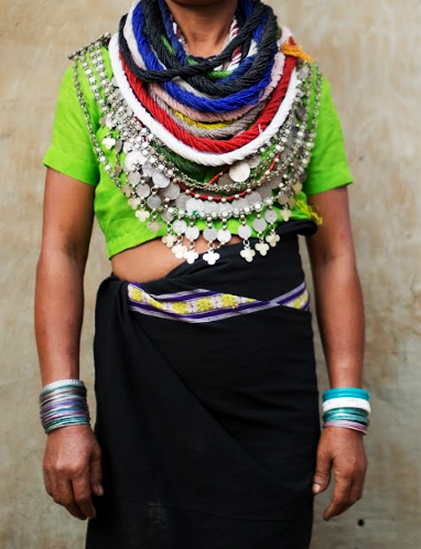Reang woman wearing a green top with multiple bead and silver coin necklaces, a black skirt with a patterned woven border and stacked bangles on both wrists