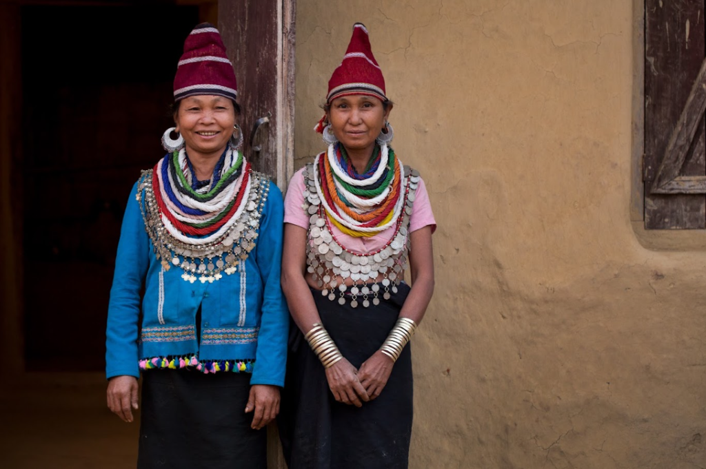 Two Reang women in the doorway of a home, wearing red caps, blue and pink tops and layered bead and silver coin necklaces with silver bangles